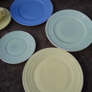 May include: Four ceramic plates in different colors: blue, light green, light green, and yellow. The plates have a raised rim and a concentric circle design.