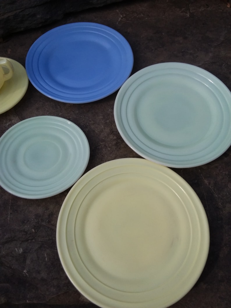 May include: Four ceramic plates in different colors: blue, light green, light green, and yellow. The plates have a raised rim and a concentric circle design.