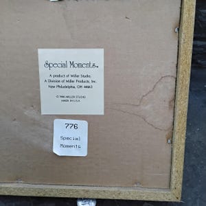 May include: The back of a framed artwork with a light brown wooden frame. A white label reads "Special Moments" and includes the Miller Studio information. Another label displays the number "776" and the words "Special Moments."