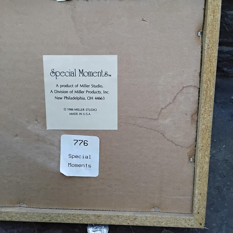 May include: The back of a framed artwork with a light brown wooden frame. A white label reads "Special Moments" and includes the Miller Studio information. Another label displays the number "776" and the words "Special Moments."
