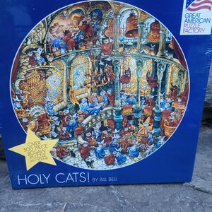May include: A square box for a 500-piece round jigsaw puzzle titled Holy Cats! by Bill Bell. The box features a circular illustration of cats in a cathedral setting, a yellow starburst label stating 50.8 cm round, and the Great American Puzzle Factory logo.