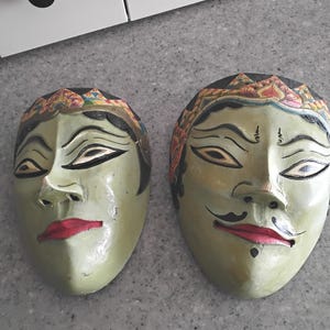 May include: Two antique, hand-painted wooden masks with green faces, red lips, and detailed headbands. The masks have a traditional design, with closed eyes and a serene expression. The masks are displayed on a gray surface.