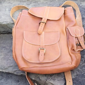 May include: A tan leather backpack with multiple pockets and a flap closure. The backpack features adjustable straps, white stitching, and brass-colored buckles. The bag has a classic, rustic design.