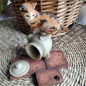 May include: A ceramic figurine of a calico cat standing on a tipped-over pot with a lid. A small brown mouse figurine is on a brick tile.
