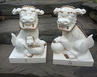May include: Two white ceramic guardian lion statues with gold-painted accents on the manes, faces, and paws. One lion holds a small cub, while the other rests a paw on a sphere. Each sits on a rectangular base with black text characters.