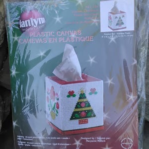 May include: A plastic canvas tissue box cover kit featuring a Christmas tree design. The kit includes 10-count plastic canvas, 4-ply acrylic yarn, and a needle. Finished size is 5" x 5" x 5.5". The packaging includes the text "PLASTIC CANVAS CANEVAS EN PLASTIQUE".