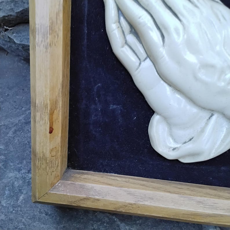 May include: A framed relief sculpture featuring clasped hands in a praying position. The hands are a light cream color, set against a dark blue background. The frame is made of light-colored wood.