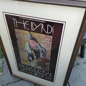 May include: Framed concert poster for The Byrds, featuring an eagle with an American flag. The poster also advertises Fleetwood Mac and John Hammond at the Fillmore West. The poster is in a brown wooden frame.