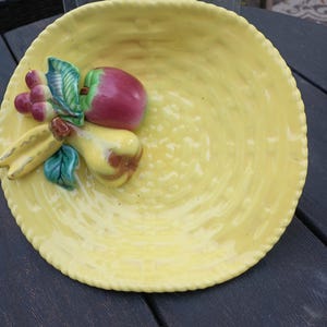 May include: A round yellow ceramic bowl with a textured basket-weave pattern on the surface. Attached to the side is a three-dimensional decorative cluster of fruit featuring a red apple, a yellow pear, purple grapes, and green leaves.