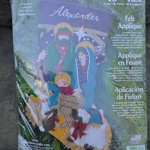 May include: Felt applique kit featuring a Nativity scene with Mary, Joseph, and baby Jesus, along with animals. The kit includes wool yarn, cotton thread, and printed felt. The package is approximately 18 inches (46 cm) long.