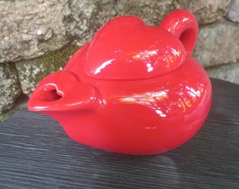 May include: A bright red ceramic teapot with a heart-shaped lid and a heart-shaped spout opening. The teapot features a smooth, glossy finish and a rounded body, with a curved handle extending from the back, showcasing a vibrant solid color.