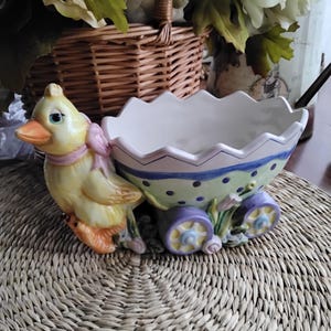 May include: A ceramic Easter chick pulling a wagon shaped like an egg. The wagon is decorated with green, purple, and white polka dots and has two wheels. The chick is yellow with a pink bow and a carrot in its beak.