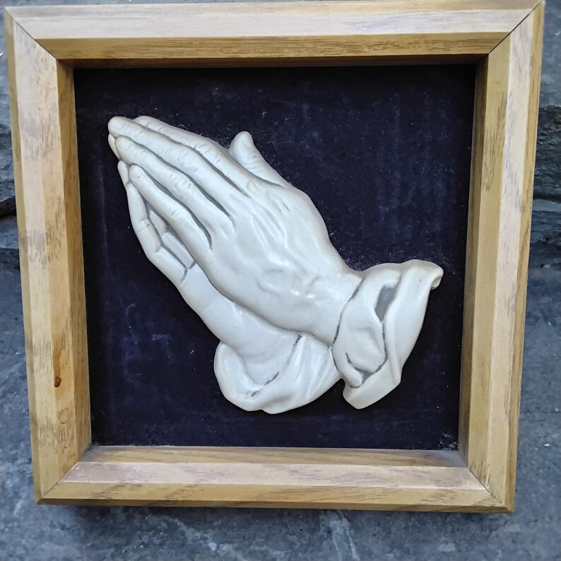 May include: A framed relief sculpture of two hands clasped in prayer. The hands are a light, off-white color and are set against a dark blue background. The frame is made of light-colored wood.