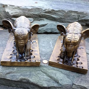 May include: A pair of bronze-toned metal elephant bookends, each featuring a textured rectangular base with a pebble-like surface. A single copper-colored penny is placed between the two figures to provide a sense of scale for the objects.