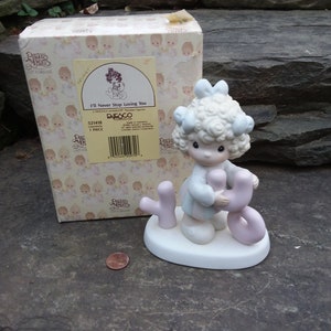 May include: A porcelain figurine of a girl with blonde curly hair wearing a blue dress and holding a pink heart with the word "You" on it. The figurine is on a white base with the word "I'll" in pink letters. The box for the figurine is white with a pink and white pattern and the words "Precious Moments" and "I'll Never Stop Loving You".