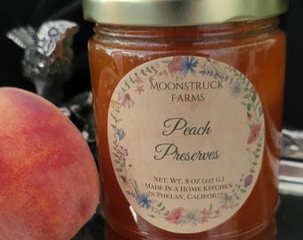 May include: A glass jar of orange peach preserves with a gold lid and a round label featuring floral illustrations. The label reads Moonstruck Farms Peach Preserves, Net. Wt. 8 oz (227 g), Made in a home kitchen in Phelan, California. A peach sits nearby.