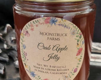 May include: A glass jar filled with dark red crab apple jelly, topped with a gold metal lid. The circular label features a floral border and reads: MOONSTRUCK FARMS, Crab Apple Jelly, NET. WT. 8 OZ (227 G.), MADE IN A HOME KITCHEN IN PHELAN, CALIFORNIA.