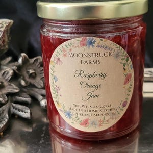 May include: A glass jar filled with red raspberry orange jam, topped with a gold metal lid. The circular label features a floral border and the text: MOONSTRUCK FARMS, Raspberry Orange Jam, NET. WT. 8 OZ. (227 G.), MADE IN A HOME KITCHEN, IN PHELAN, CALIFORN.
