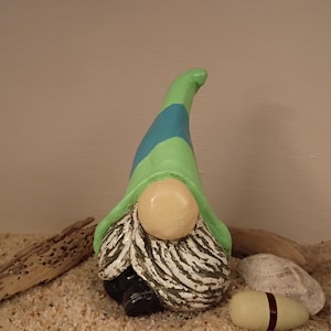 May include: A ceramic gnome figurine with a green and blue hat, a tan face, and a white and gray beard. The gnome is set on a bed of sand with seashells and driftwood.