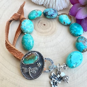 May include: A bracelet featuring smooth, oval turquoise beads with natural dark veining, strung on a brown leather cord. It includes a circular silver pendant with a dragonfly engraving, a small turquoise inlay, and dangling silver flower charms.