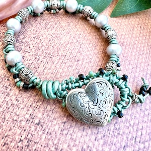 May include: A handcrafted bracelet with a silver heart pendant, adorned with an ornate pattern. The bracelet combines light blue leather, white pearls, and silver beads, offering a distinctive and fashionable accessory.