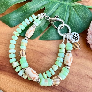 May include: A double-strand bracelet featuring light green beads and decorative beige and brown beads. The bracelet has a silver clasp and a charm with a tree design. The bracelet is displayed on a wooden surface.