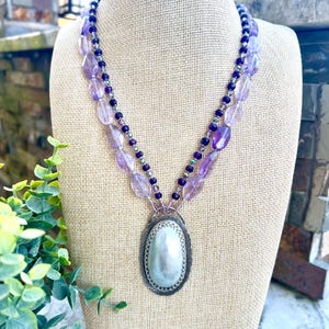 May include: A layered necklace with purple and clear beads, and a large oval pendant. The pendant has a silver-colored frame and a pearlescent center. The necklace is displayed on a beige mannequin.