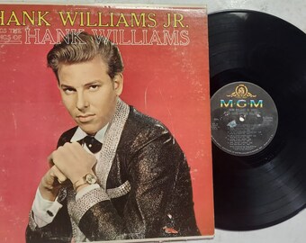 May include: A vinyl record and its square cardboard cover featuring a portrait of Hank Williams Jr. The red cover displays the text HANK WILLIAMS JR. SINGS THE SONGS OF HANK WILLIAMS and an MGM logo. The black vinyl record has a central MGM label.