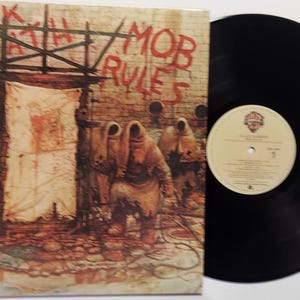May include: Black Sabbath vinyl record album cover art featuring a dark, gritty, and apocalyptic scene with hooded figures in a dilapidated building. The album title "Mob Rules" is written in red spray paint on the wall.
