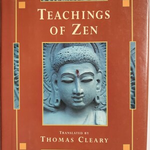 May include: A hardcover book titled "Teachings of Zen" with a terracotta-colored cover. The cover features a serene, light blue sculpture of a face with closed eyes. The text "Translated by Thomas Cleary" is also visible.