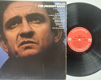 Pode incluir: Uma capa de álbum de vinil com um retrato de close-up de Johnny Cash. O título do álbum é "Hello, I'm Johnny Cash" e a gravadora é Columbia. A capa do álbum é azul claro com um fundo azul escuro.