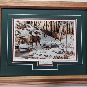 May include: Framed print of a buck standing in a snowy stream. The artwork, titled "River Bottom Buck," features a detailed depiction of the deer in a winter forest scene, framed in a wooden frame with a green mat.