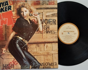 Pode incluir: Uma capa de álbum de vinil com uma mulher cantando em um microfone. O álbum é intitulado "TNT" e apresenta o nome da artista "Tanya Tucker" em letras vermelhas grandes. O fundo é uma parede de madeira com as palavras "Danger High Explosives" escritas nela.
