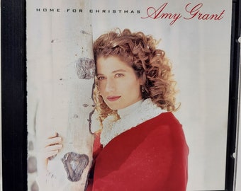 May include: CD case featuring Amy Grant, with the text "HOME FOR CHRISTMAS" and "Amy Grant" in red. The cover shows a woman with curly hair, wearing a red shawl and white collar, leaning against a tree trunk.
