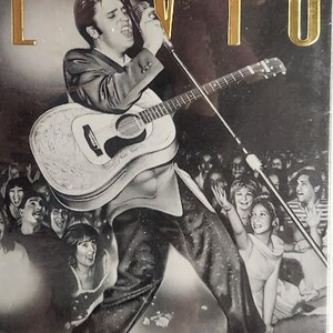 May include: A black and white photo of Elvis Presley performing on stage with a guitar. The photo is from a concert and shows Elvis singing into a microphone with a crowd of fans in the background. The text "THE GREAT PERFORMANCES ELVIS CENTER STAGE" is printed on the image.