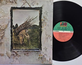 May include: A Led Zeppelin IV vinyl record and its album cover. The cover features a framed image of a person carrying a large bundle of sticks on their back against a field. The record label is green and orange with Atlantic branding.