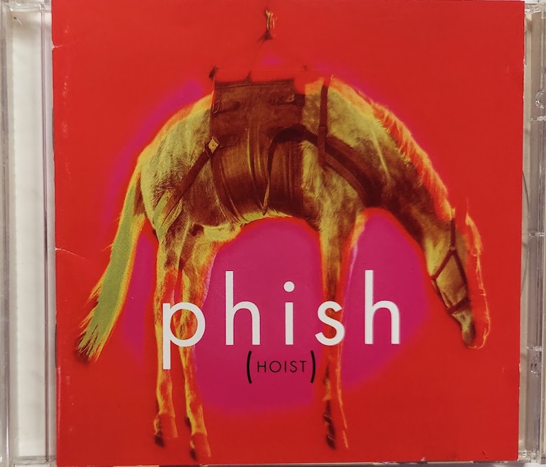 May include: A red CD case with a white horse on the front. The horse is upside down and has a saddle on its back. The text "phish (HOIST)" is printed in white on the front of the case.