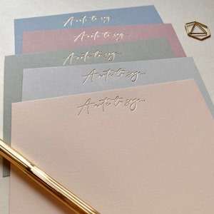 May include: A collection of pastel-coloured writing paper, each sheet embossed with the text "A note to say...". The paper is in shades of blue, pink, and grey. A gold pen and a geometric paperclip are also present.