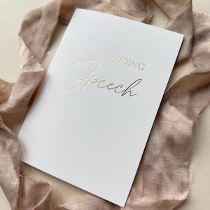 May include: A white card with gold foil lettering that reads "WEDDING Speech". The card is angled and rests on a soft, blush-coloured ribbon. The overall aesthetic is elegant and minimalist.
