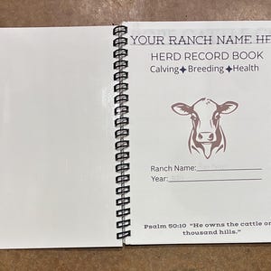 May include: A white spiral-bound herd record book featuring a brown cow illustration and text that reads: YOUR RANCH NAME HERE, HERD RECORD BOOK, Calving, Breeding, Health, Ranch Name, Year, and Psalm 50:10 He owns the cattle on a thousand hills.