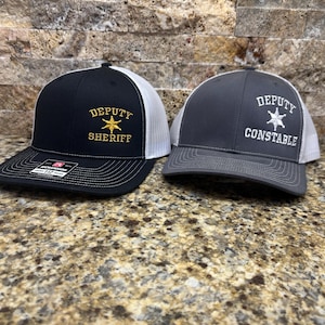 May include: Two black and gray trucker hats with white mesh back panels. The hat on the left has the text "DEPUTY SHERIFF" embroidered in gold with a gold star. The hat on the right has the text "DEPUTY CONSTABLE" embroidered in white with a white star.