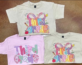 May include: Three short-sleeved t-shirts in beige and light pink. Two beige shirts feature a graphic with a large pink bow, school supplies, and the text Third Grade or First Grade. One pink shirt features the text Third Grade in floral patterns.