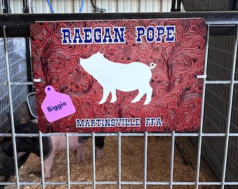 May include: A rectangular sign with a red and black floral tooled leather pattern featuring a white pig silhouette in the center. The text RAEGAN POPE is printed at the top, MARTINSVILLE FFA at the bottom, and a pink ear tag labeled Biggie is attached.
