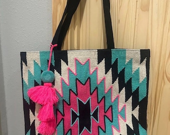 May include: A rectangular tote bag featuring a woven geometric pattern in black, white, turquoise, and hot pink. The bag has two black shoulder straps and a decorative hanging tassel accessory in turquoise and hot pink attached to the side.