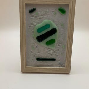 Peut inclure: Œuvre d'art abstraite dans un cadre en bois clair. L'œuvre présente un motif vert clair, noir et vert foncé sur un fond blanc texturé. Le cadre est rectangulaire et en bois de couleur claire.