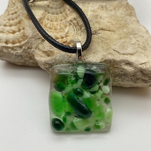 Peut inclure: Un collier avec un pendentif carré et un cordon noir. Le pendentif transparent contient des morceaux de verre vert de différentes nuances. Le collier a un fermoir argenté.