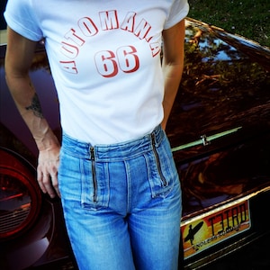 May include: A woman wearing a white t-shirt with the text "AUTOMANIA 66" in red. She is wearing blue jeans with zipper details and is standing next to a vintage car with a license plate that reads "73180 ENDLESS SUMMER".