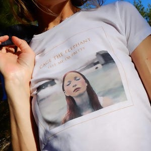May include: White t-shirt with the text "CAGE THE ELEPHANT TELL ME I'M PRETTY" in a gold color. The shirt features a framed image of a woman with red hair and closed eyes. The shirt is short-sleeved.