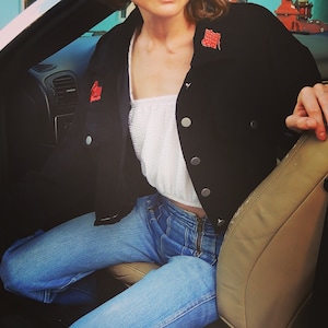 Puede incluir: Una mujer con una chaqueta vaquera negra con parches bordados rojos, un top blanco corto y unos vaqueros azules. Está sentada en un coche.