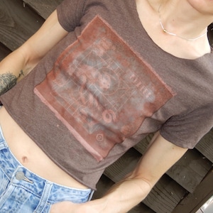 Puede incluir: Un top corto marrón con un diseño gráfico cuadrado en tonos rojos y marrones. Se combina con vaqueros de mezclilla azul claro. La imagen presenta un estilo casual y cotidiano.